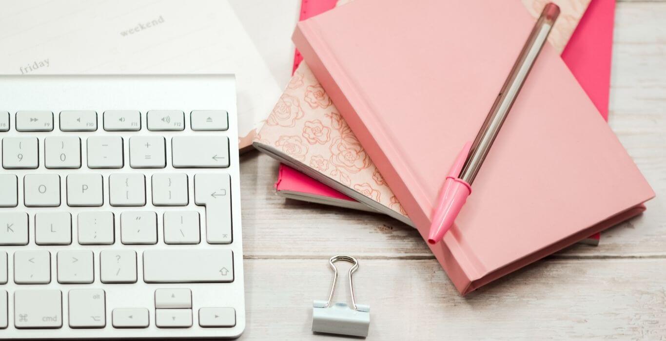 white keyboard and stack on pink notebooks on a white wooden background