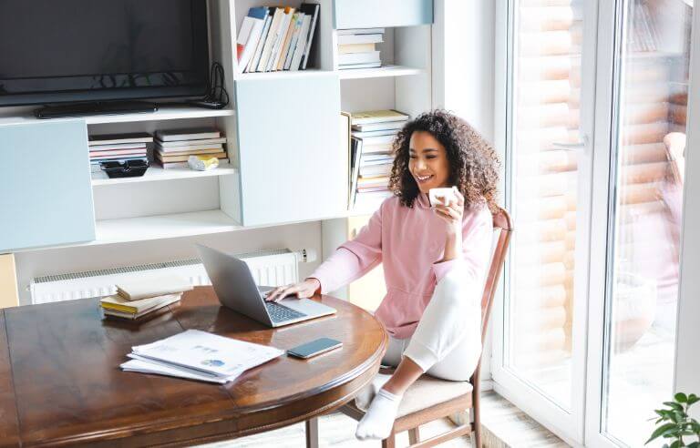 Woman sitting at dining room table, holding a cup of coffee while looking at web hosting options on her laptop