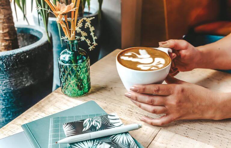 hands holding a cup of coffee on a cafe table to notebooks stacked near by