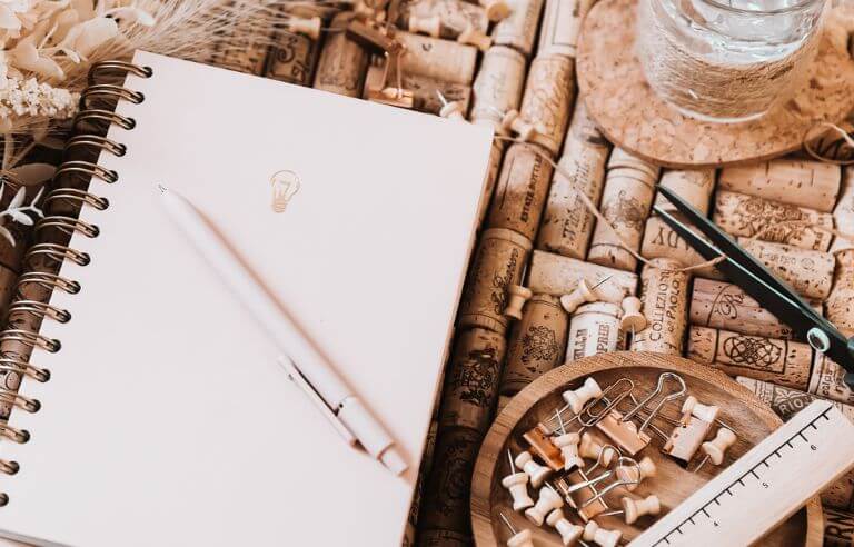 neutral colored organizer, pen, paperclips and pushpins in wooden bowl on a corkboard backdrop