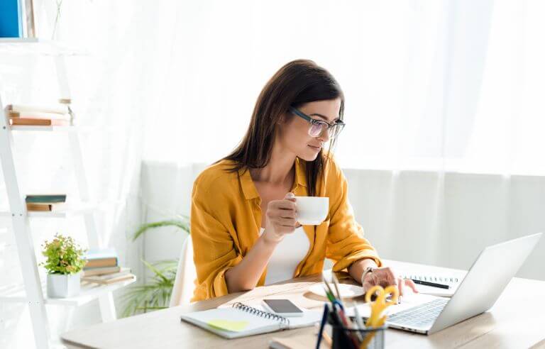 woman in yellow shirt holding a cup of coffee while working on laptop
