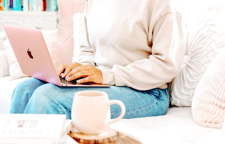 woman sitting on a white couch working on her website's Alt Text on her rose colored laptop, coffee mug on table in front of her